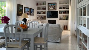 Een woonkamer met een eettafel, stoelen, een bank, een televisie en rekken met diverse spullen.