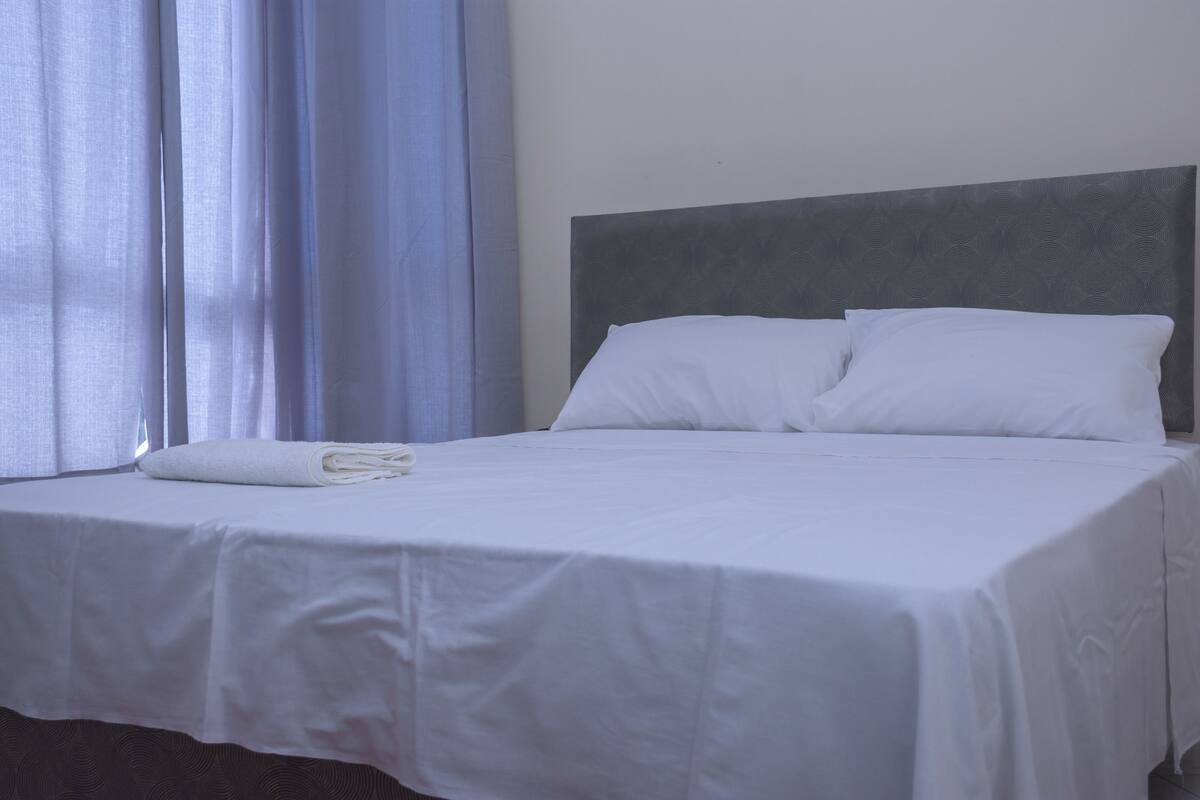 A neatly made bed with white linens and pillows, a grey headboard, and a window with blue curtains.