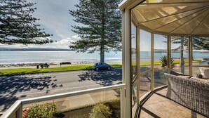 Un balcon fermé par des baies vitrées, offrant une vue sur la plage, une voiture garée dans la rue et un grand arbre.