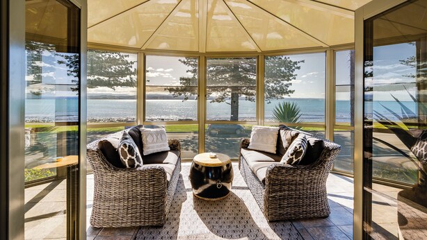 Une véranda avec une structure en verre, des meubles en osier et une vue sur la mer.