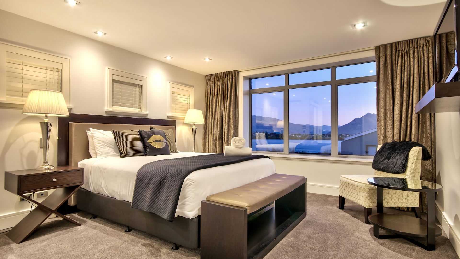 Une chambre d’hôtel moderne dotée d’un grand lit, de deux tables de chevet, d’un banc, d’une chaise et d’une grande fenêtre donnant sur les montagnes.