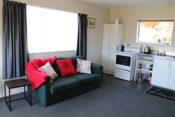 A compact living space with a green sofa, a small table, a kitchenette, and a window with curtains. - Somers holiday cottage (Mt Somers)