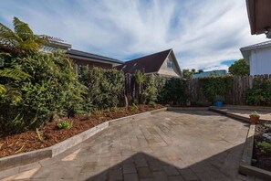 Une terrasse dallée, surmontée d’une structure de toit inclinée, entourée d’une végétation luxuriante et d’une clôture en bois.