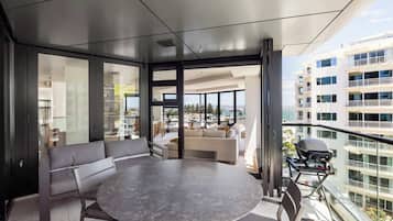 A modern balcony with a round table and seating, large windows offering a city view, and a grill on the right.