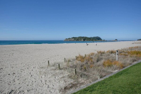 A sandy beach with a grassy area and a wooden fence. - Luxury Ocean Views - Heated Pool, Main Beach & Gym (Mount Maunganui)