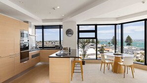 Een moderne keuken met een centraal eiland, grote ramen met uitzicht op stad en zee, en een eethoek met stoelen en een tafel.