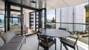 Een moderne woonkamer met een bank, stoelen en een tafel, grote ramen met uitzicht op een stad en water, en een balkon met uitzicht op een bouwplaats.