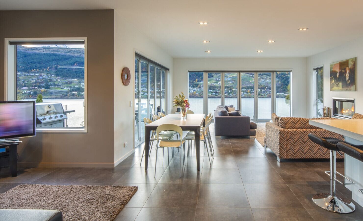 A modern living room with a dining area, large windows, a fireplace, and a TV.