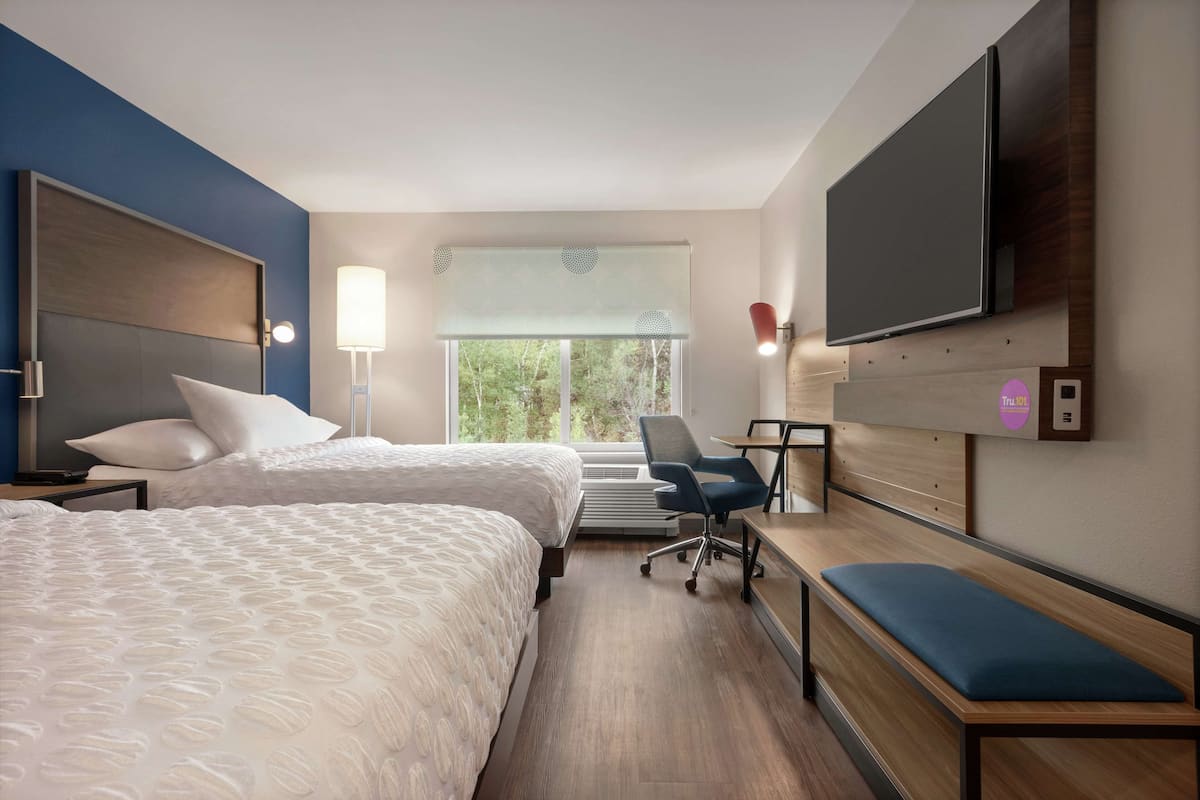 Habitación de hotel con dos camas, un escritorio, una banca, un televisor y una ventana con vistas a árboles.