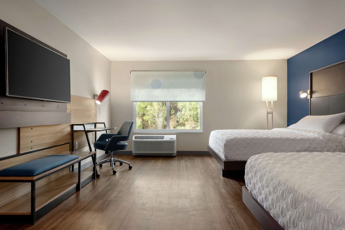Una habitación de hotel con cama, escritorio, silla, televisión y ventana con persianas.
