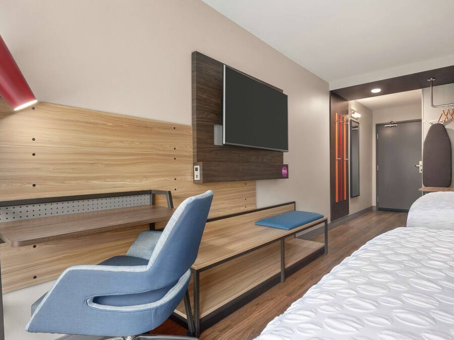 A modern hotel room with a wooden headboard, a flat-screen TV mounted on the wall, a desk with a chair, and a bed with a patterned duvet.