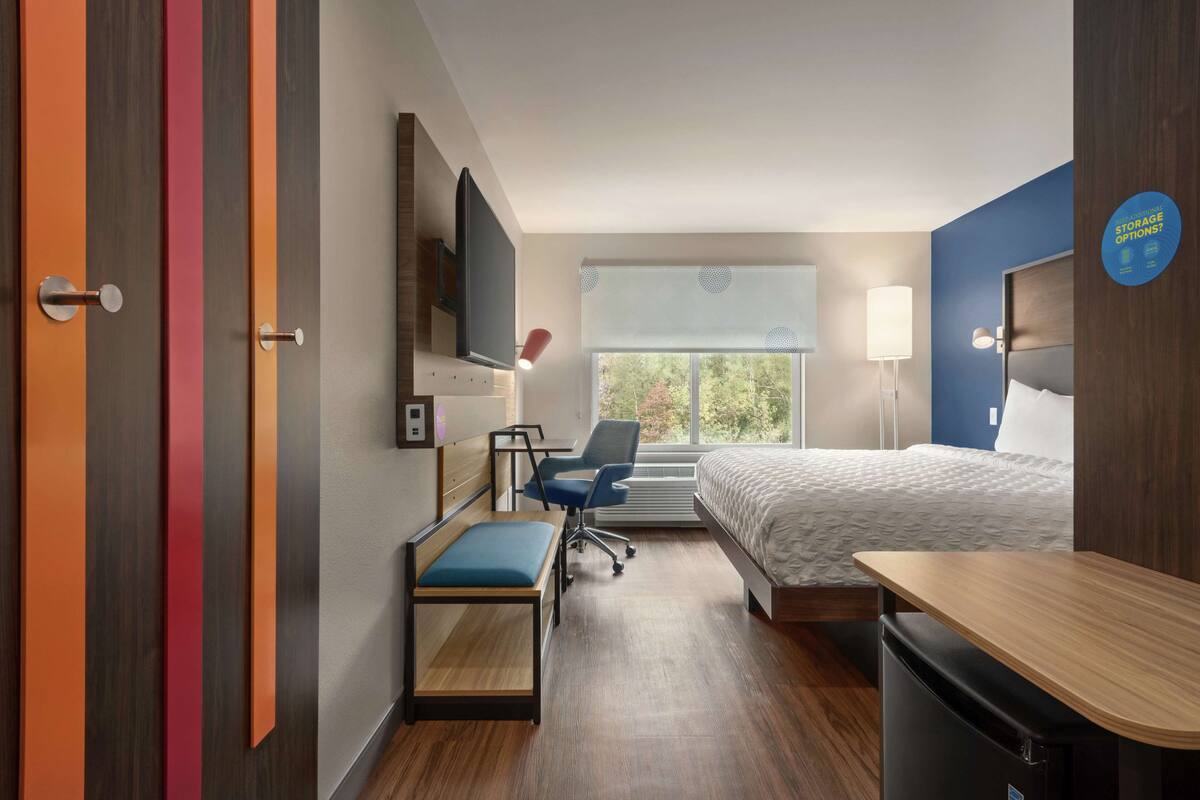 Habitación de hotel con cama, escritorio, silla, televisor y ventana con vista a árboles.