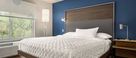 A modern bedroom with a large bed, a nightstand, a desk, and a window with a view of trees.
