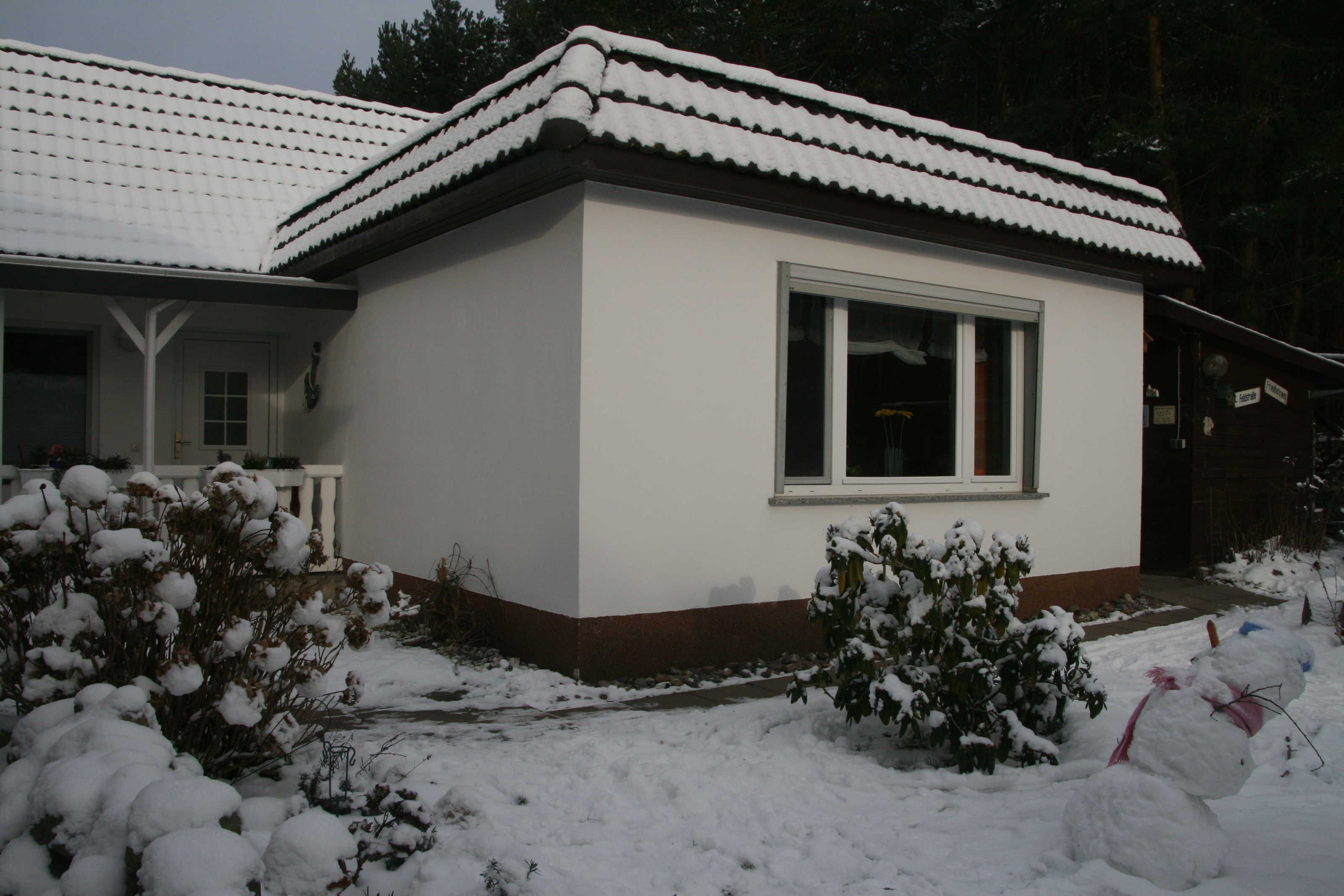 Ein kleines, eingeschossiges Gebäude mit weissem Anstrich und einem Ziegeldach, umgeben von schneebedecktem Boden und Pflanzen.