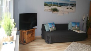 A living room with a TV, a sofa with cushions, a wall with beach-themed artwork, and a plant. - Vacation home at the edge of the forest Biendorf near Kühlungsborn - 2-room vacation home (70m², max. (Biendorf)