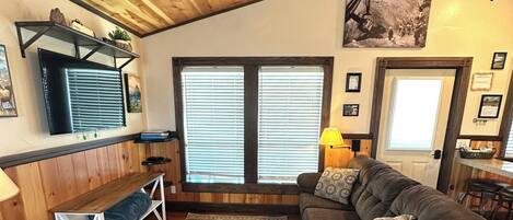 A cozy living room with a wooden ceiling, a large window with blinds, a sofa, a coffee table, and a TV on a shelf.