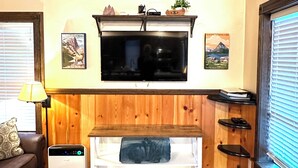 Una sala de estar acogedora con techo de madera, un televisor de pantalla plana sobre un banco de madera, un calentador pequeño y una ventana con persianas.