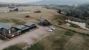 Vista aérea de una propiedad rural con varias construcciones, incluyendo una casa grande con porche y una cabaña más pequeña. Hay varios coches aparcados frente a los edificios.