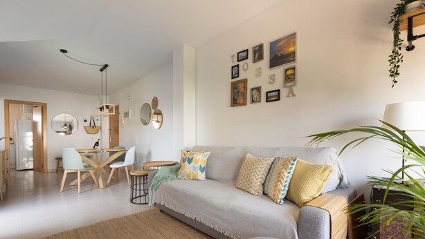 A modern living room with a grey sofa, a dining area with a wooden table and chairs, and a kitchen visible in the background.