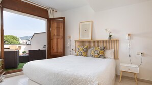 A bedroom with a large bed, a wooden headboard, a bedside table, a small shelf with decorative items, and a view of the outdoors.