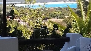 Un balcón con un sofá de mimbre blanco y una mesita pequeña, con vistas a un paisaje costero con vegetación y una vista distante del mar.