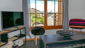 A room with a television on a stand, a chair, a small table with a bowl, and a view of a building and greenery through a large window. - Ferme de Sandra F2 by Interhome (Nendaz)