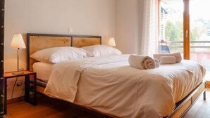 A bedroom with a wooden bed frame, white bedding, a bedside table with a lamp, and a view of the outdoors through a glass door. - Ferme de Sandra F2 by Interhome (Nendaz)