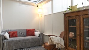 Una habitación acogedora con un sofá gris, cojines rojos y blancos, un tapete a rayas, un aparador de madera con puertas de cristal y un radiador.