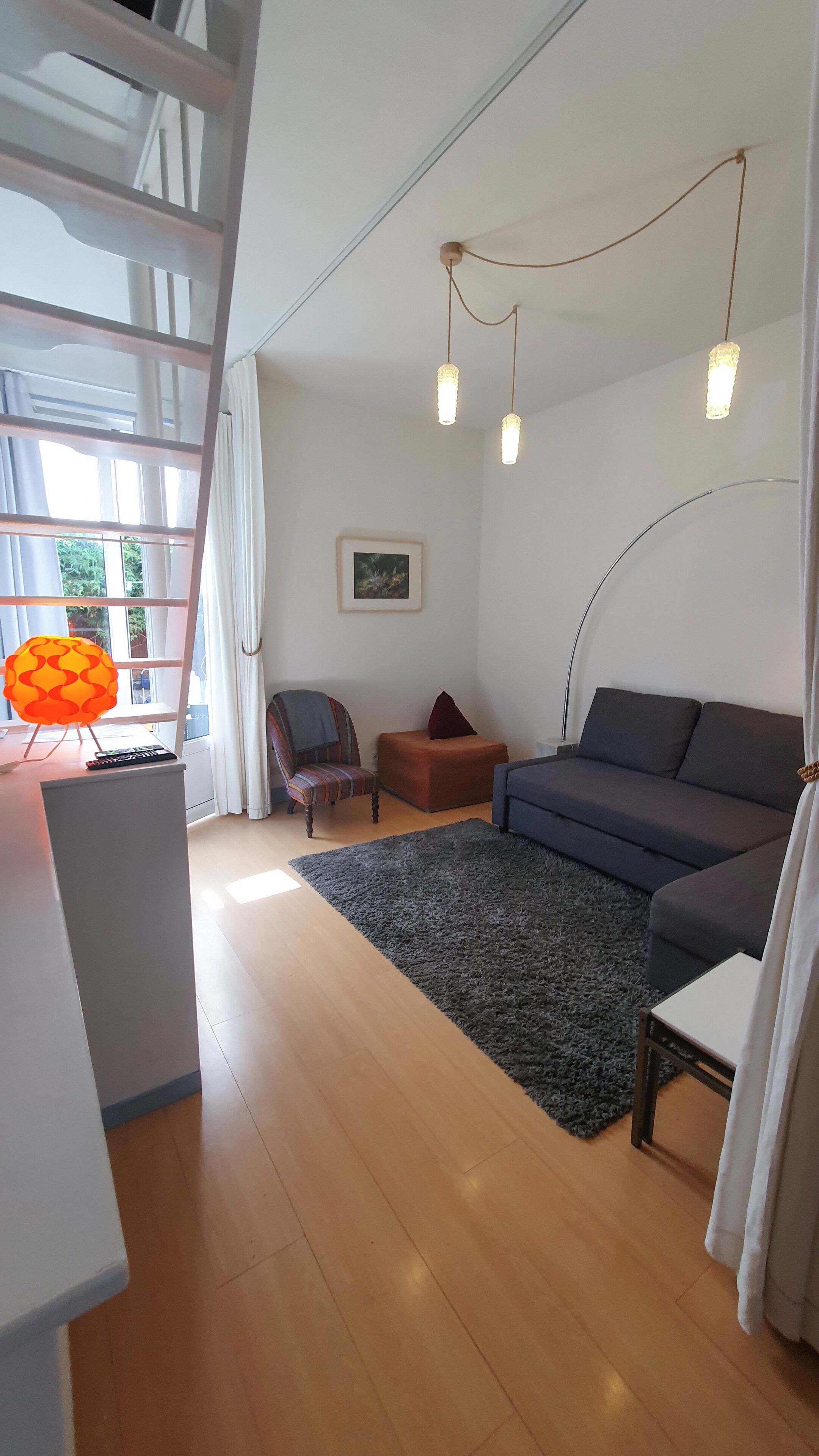 Un salon moderne avec un canapé, un fauteuil et une petite table. On y trouve un escalier avec une rampe, une fenêtre avec des rideaux et un luminaire suspendu.