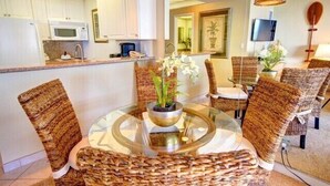 A modern kitchen with white cabinets, a microwave, and a dining area with wicker chairs and a glass table. - Tower 1 South facing. Beautiful Ocean, Lanai , koi pond and courtyard views (Lahaina)