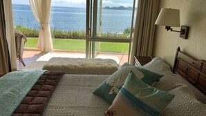 A bedroom with a large window offering a view of the sea. There is a bed with a patterned bedspread and pillows, a bedside table, and a lamp.
