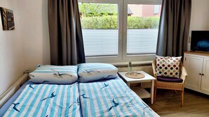A room with a bed, a chair, a small table, a television, and a window with curtains.