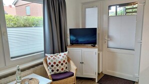 A room with a television on a cabinet, a wicker chair with a polka dot cushion, a small table with a bottle and glass, and a window with curtains.