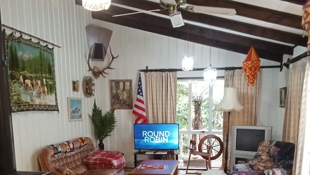 Una sala de estar acogedora con un televisor mostrando "ROUND ROBIN", una chimenea, un ventilador de techo y un gran ventanal con cortinas.
