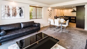 A modern living room with a black leather sofa, a glass coffee table, and a dining area with white chairs. A large painting of horses is on the wall, and there is a kitchen in the background. - 3 LEVEL LAKESIDE LUXURY WITH MAGNIFICENT LAKE & MOUNTAIN VIEWS POTENTIAL CARHIRE (Frankton)