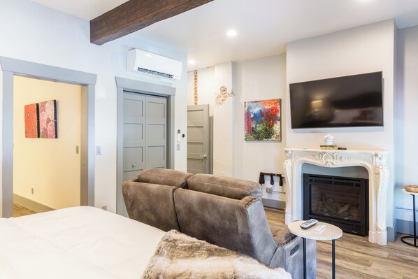 A modern hotel room with a large bed, a sofa, a fireplace, and a TV mounted on the wall.