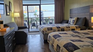 Habitación de hotel con dos camas, un escritorio con lámpara, una silla y vistas a la ciudad a través de amplias ventanas.