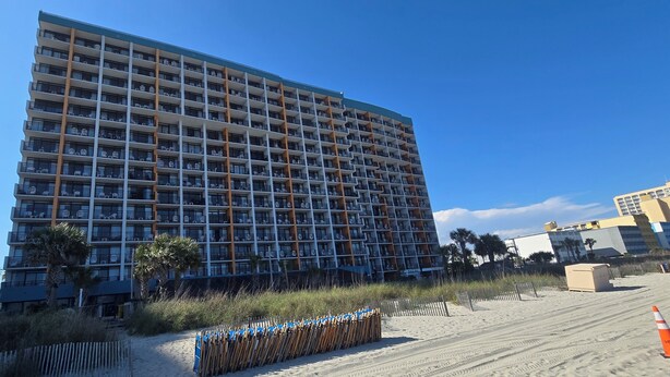Un hotel de gran altura con varios balcones, situado cerca de una playa con arena en primer plano y un cielo azul despejado.