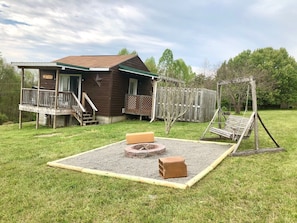 A wooden cabin with a porch, a swing set, and a fire pit in the yard.