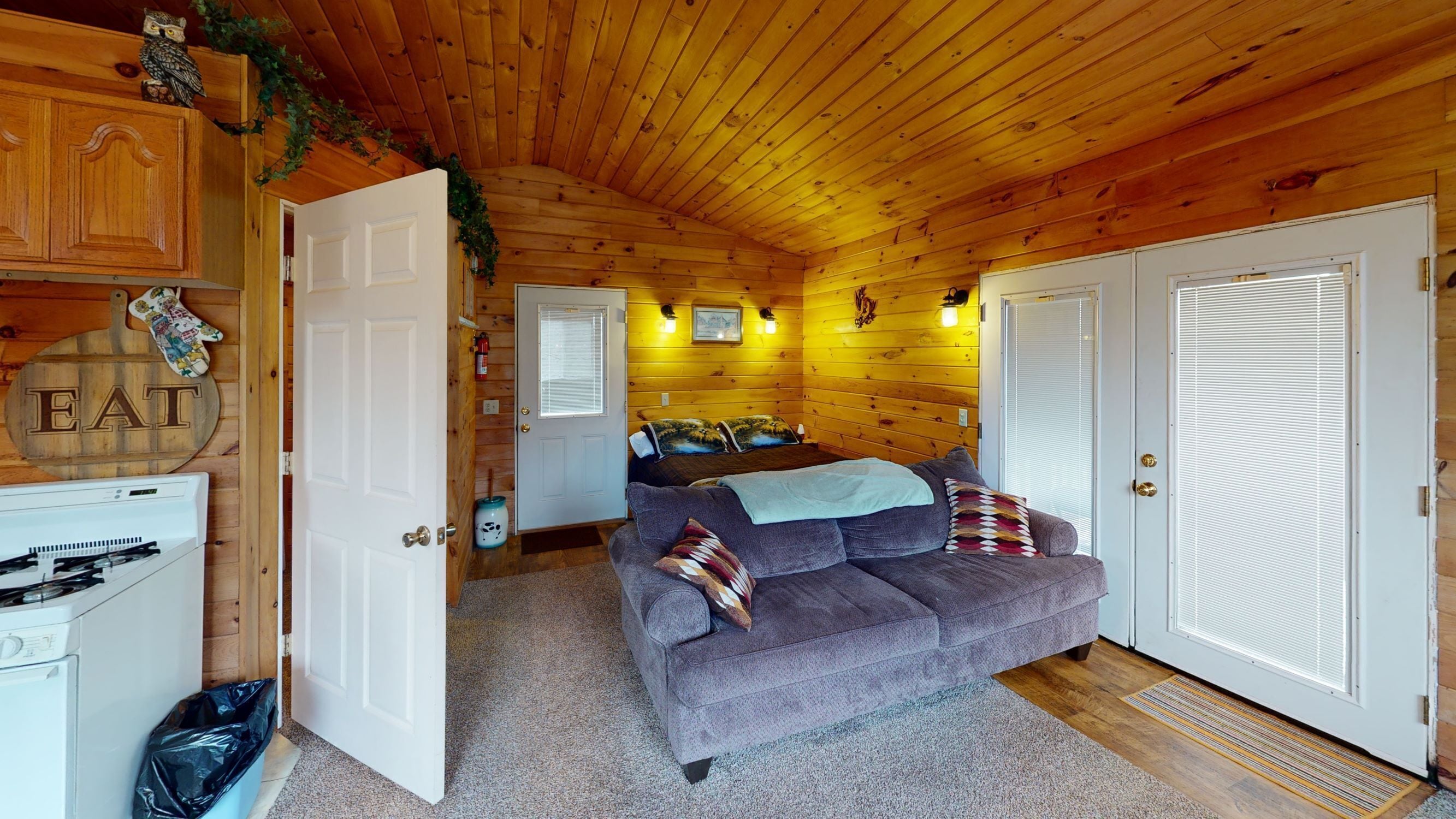 A cozy cabin interior with a wooden ceiling, a gray sofa with pillows, a dining area with a wooden cabinet and a sign that reads 'EAT', and a white door with a window.