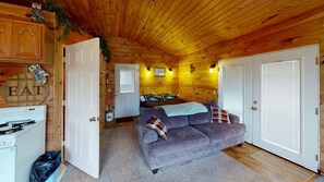 Ein gemütliches Blockhaushaus-Interieur mit Holzbalkendecke, einem grauen Sofa mit gemusterten Kissen, einem Bett mit Kopfstütze im Waldstil und einer Küchenzeile mit Herd und Mülleimer.