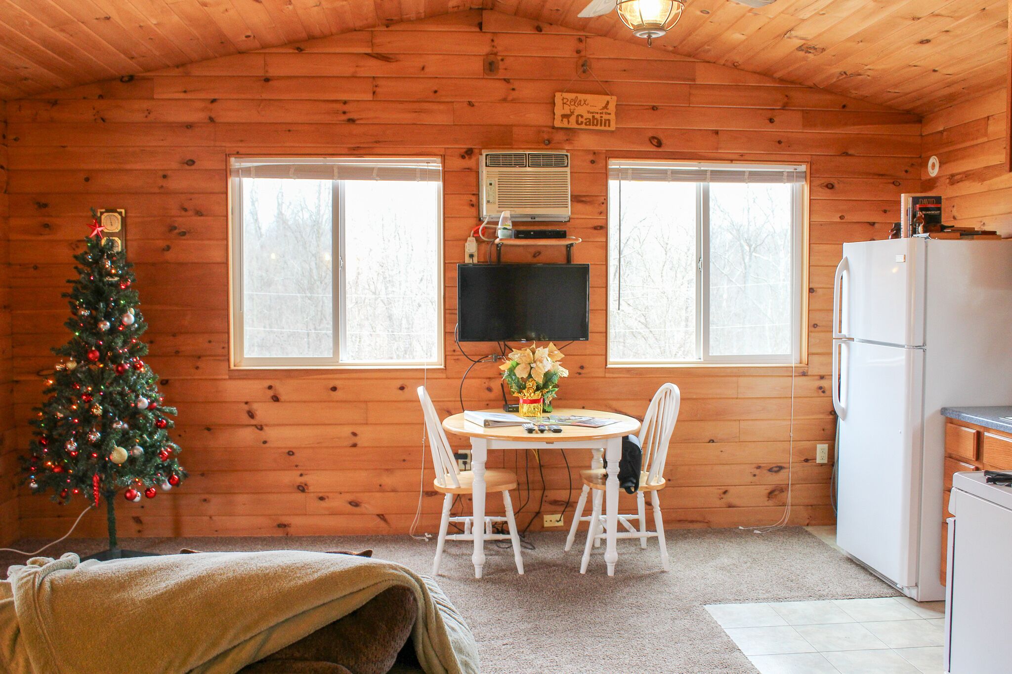 L’intérieur chaleureux d’un chalet, avec un décor en bois, une table à manger et des chaises, un sapin de Noël, un téléviseur et un réfrigérateur.