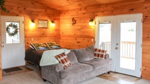 Habitación tipo cabaña con techo de madera, un sofá gris con almohadones, dos puertas y una ventana con vistas.