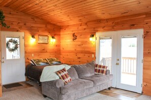 Ein gemütliches Blockhausszimmer mit Holzbalkendecke, einem grauen Sofa mit Kissen, zwei Türen und einem Fenster mit Aussicht.