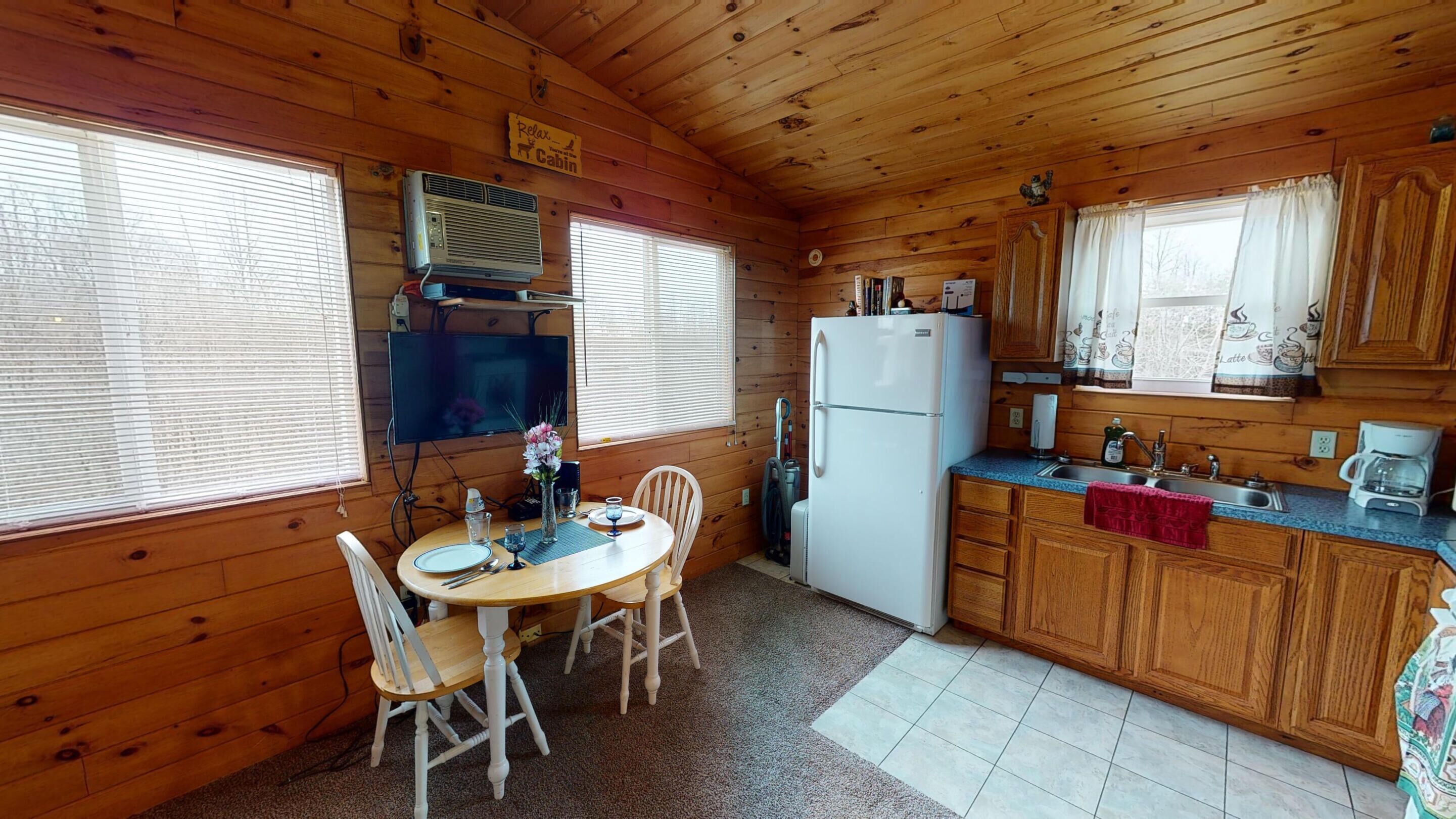 A cabin-style kitchen with wooden walls, a dining table with chairs, a refrigerator, a coffee maker, and a television.
