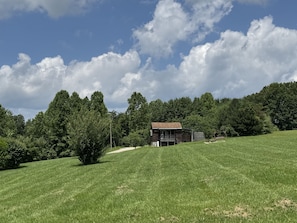 Eine kleine Holzhütte auf einem grasbewachsen Hügel, mit blauem Himmel und dichtem Wald im Hintergrund.