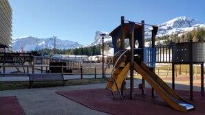 Ein Spielplatz mit Rutsche und Klettergerüst, Bänken und Blick auf verschneite Berge.
