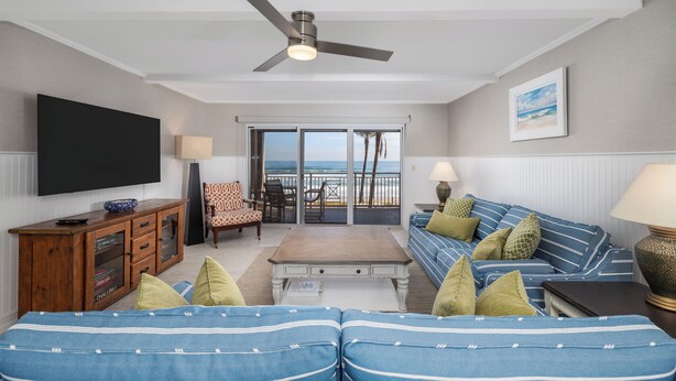 A living room with a TV, wooden cabinet, blue-striped sofa, and a ceiling fan. - Bring swim suit and start your beach holiday in this well appointed 2 bedroom 2 bath 3rd floor condo (New Smyrna Beach)