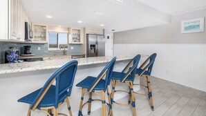 A modern kitchen with white cabinets, a stainless steel refrigerator, and a marble countertop. There are three blue wicker bar stools. - Bring swim suit and start your beach holiday in this well appointed 2 bedroom 2 bath 3rd floor condo (New Smyrna Beach)