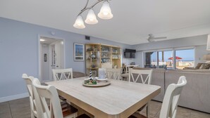 A dining area with a table and chairs, a built-in bookshelf, and a view of the beach through large windows. - Centrally located ground floor two bedroom, walk right out to pools. (New Smyrna Beach)